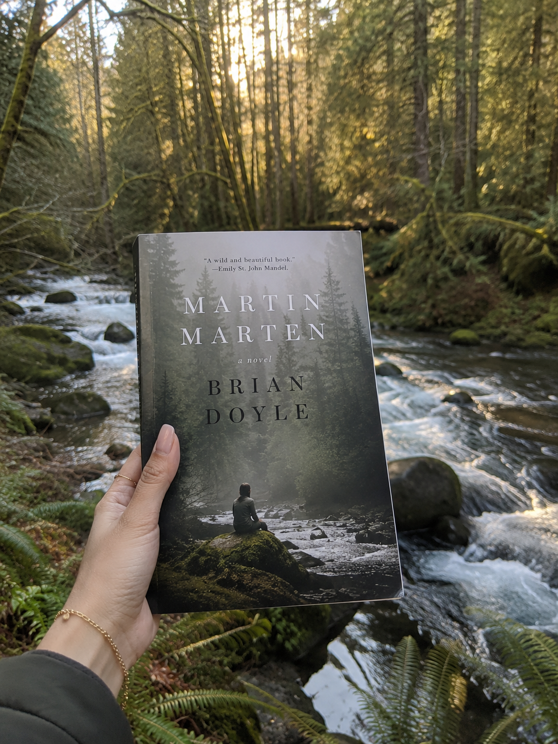 Hand holding copy of Martin Marten in front of a river in the forest